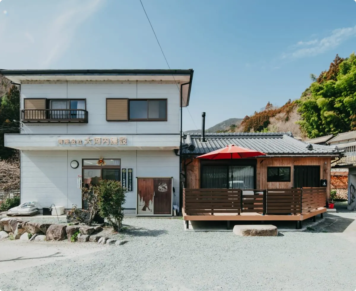 大河内建設の事務所外観。左側は白い2階建ての建物、右側にはウッドデッキと赤いパラソルのある木造の建物。背後には山と緑が広がる。